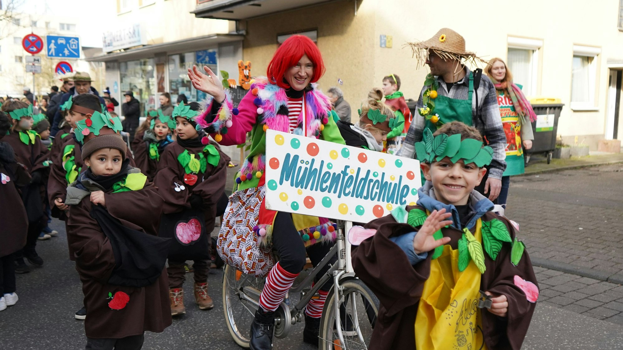 Das Foto zeigt Kinder, die als Pflanzen verkleidet sind und eine Lehrerin auf dem Fahrrad mit einem Schild, auf dem steht „Mühlenfeldschule“.