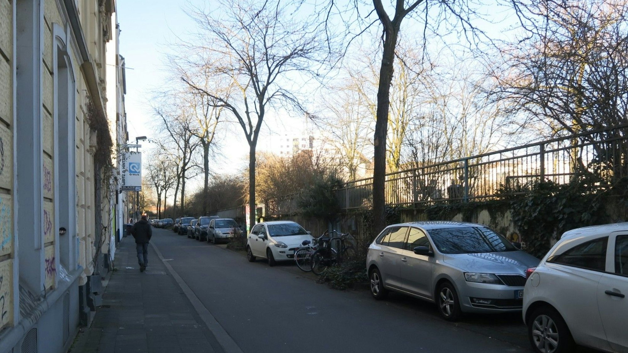 Eine schmale Straße mit geparkten Autos auf einer und einem schmalen Bürgersteig auf der anderen Seite ist zu sehen.