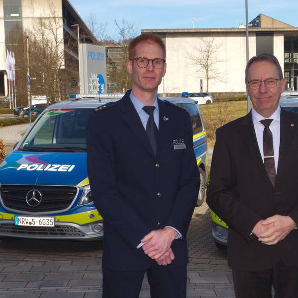 Zwei Männer stehen vor einer Polizeiwache in Gummersbach. Im Hintergrund sind zwei Polizeiautos zu sehen. Einer der Männer trägt eine Polizeiuniform.