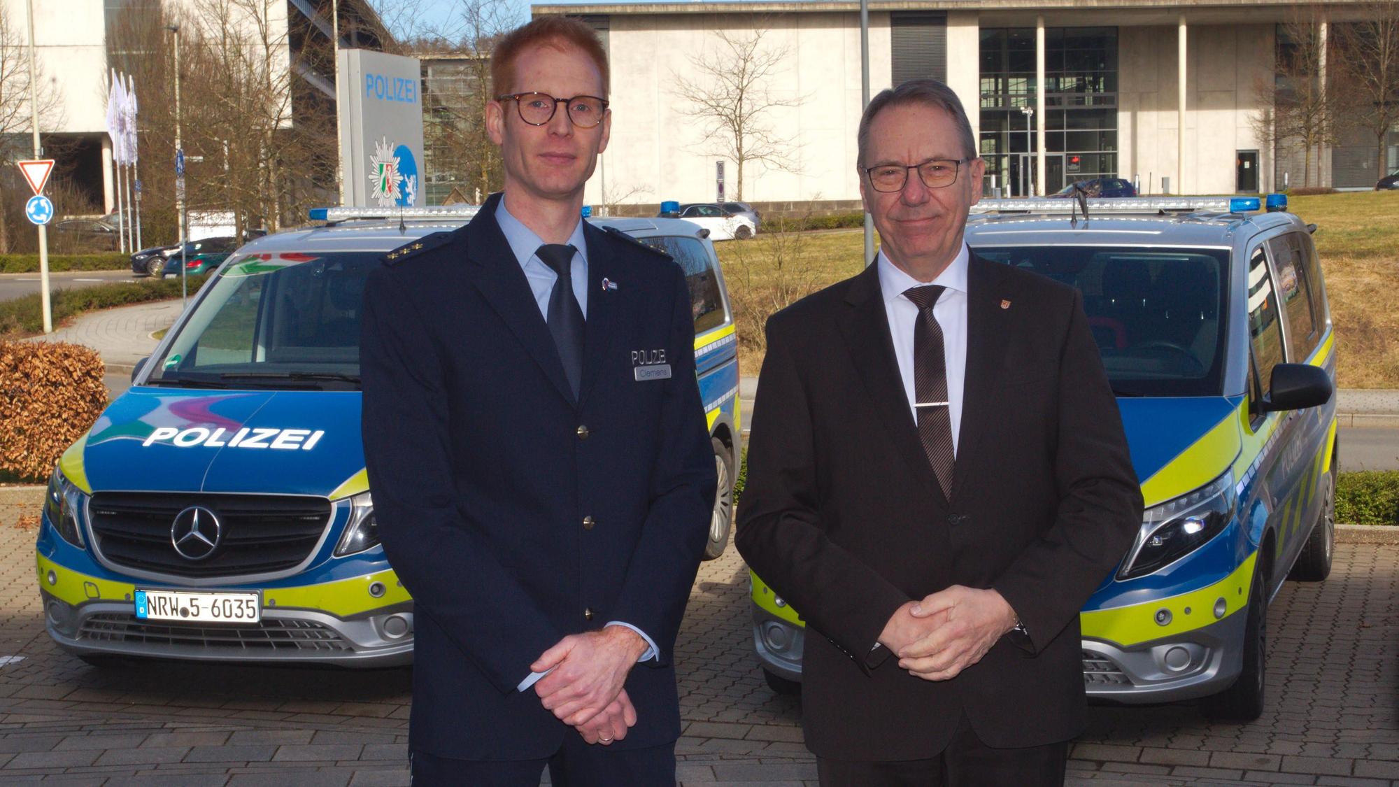 Zwei Männer stehen vor einer Polizeiwache in Gummersbach. Im Hintergrund sind zwei Polizeiautos zu sehen. Einer der Männer trägt eine Polizeiuniform.
