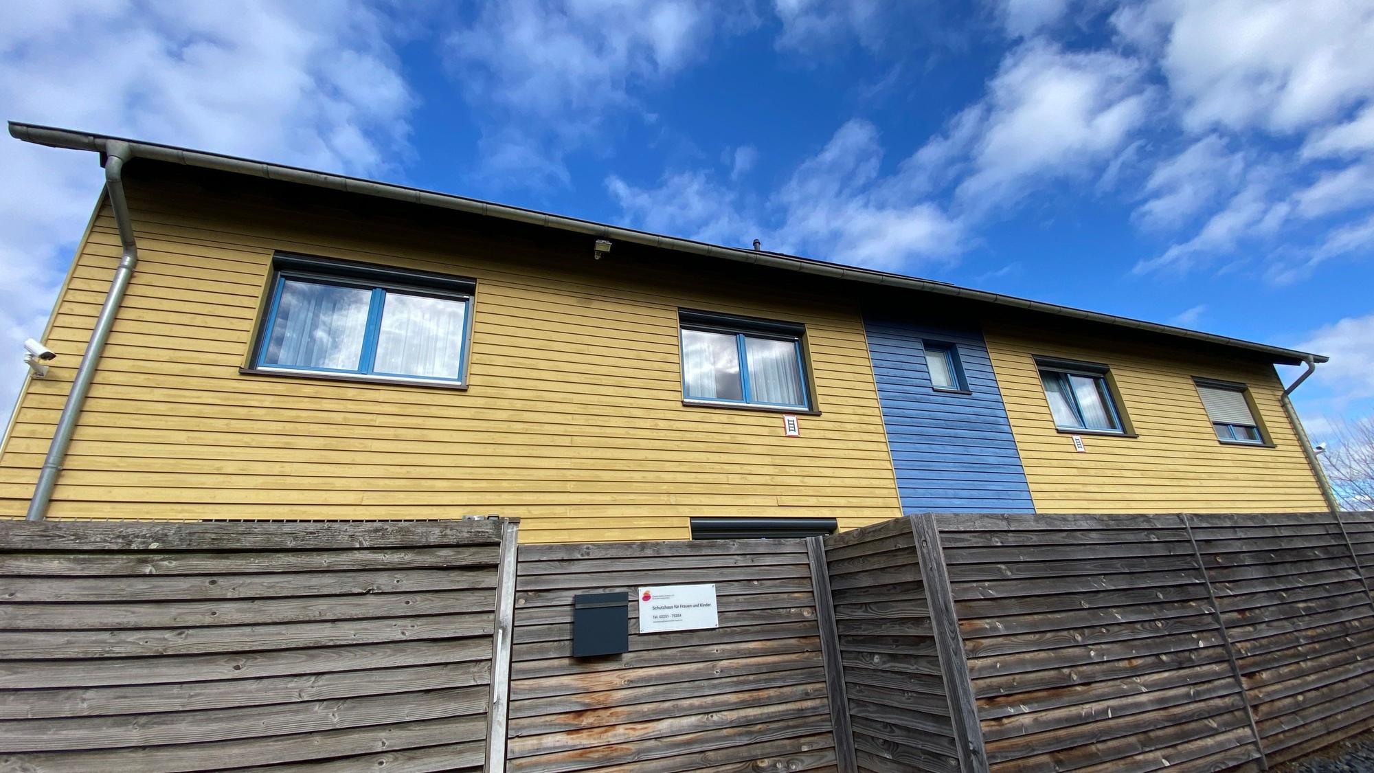 Das Gebäude in den Farben Gelb und Blau besteht aus Holz, eine Holzwand ist davor angebracht.