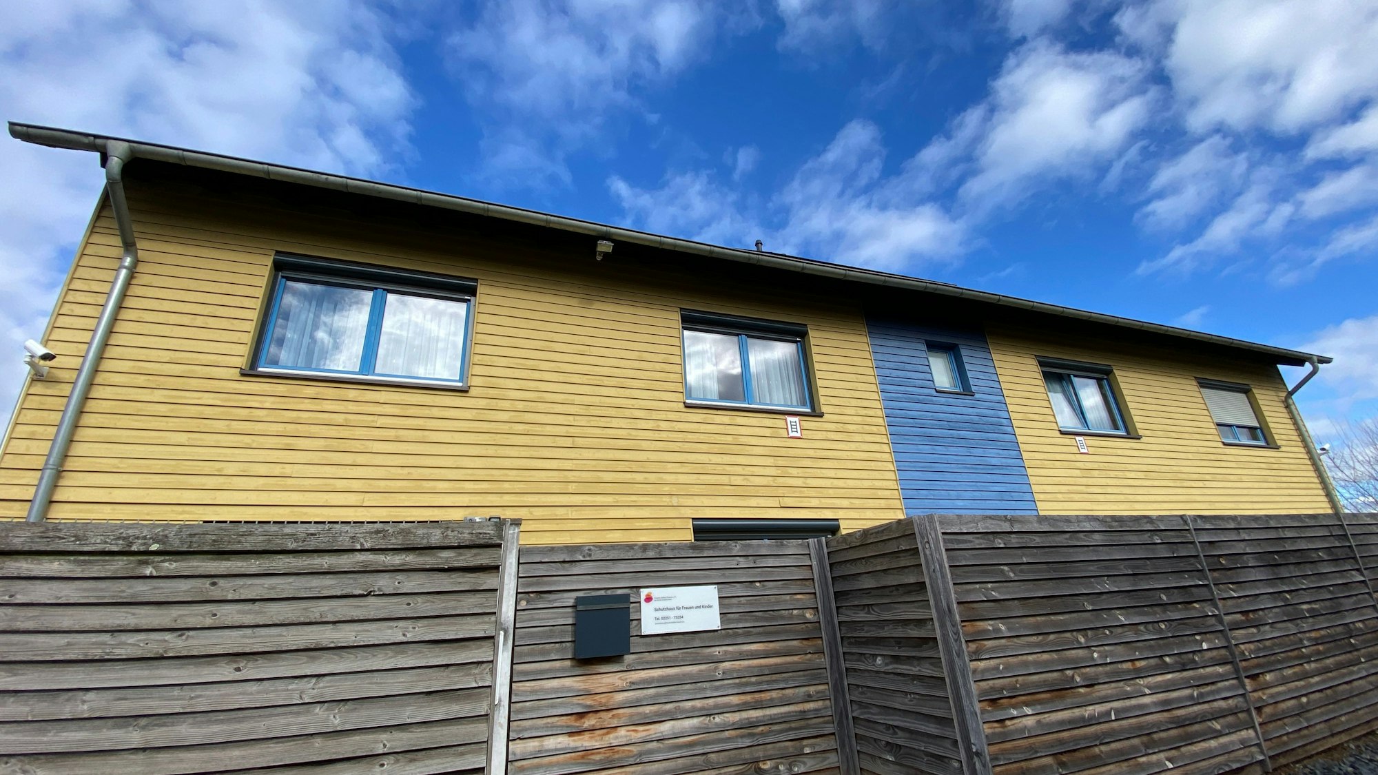 Das Gebäude in den Farben Gelb und Blau besteht aus Holz, eine Holzwand ist davor angebracht.