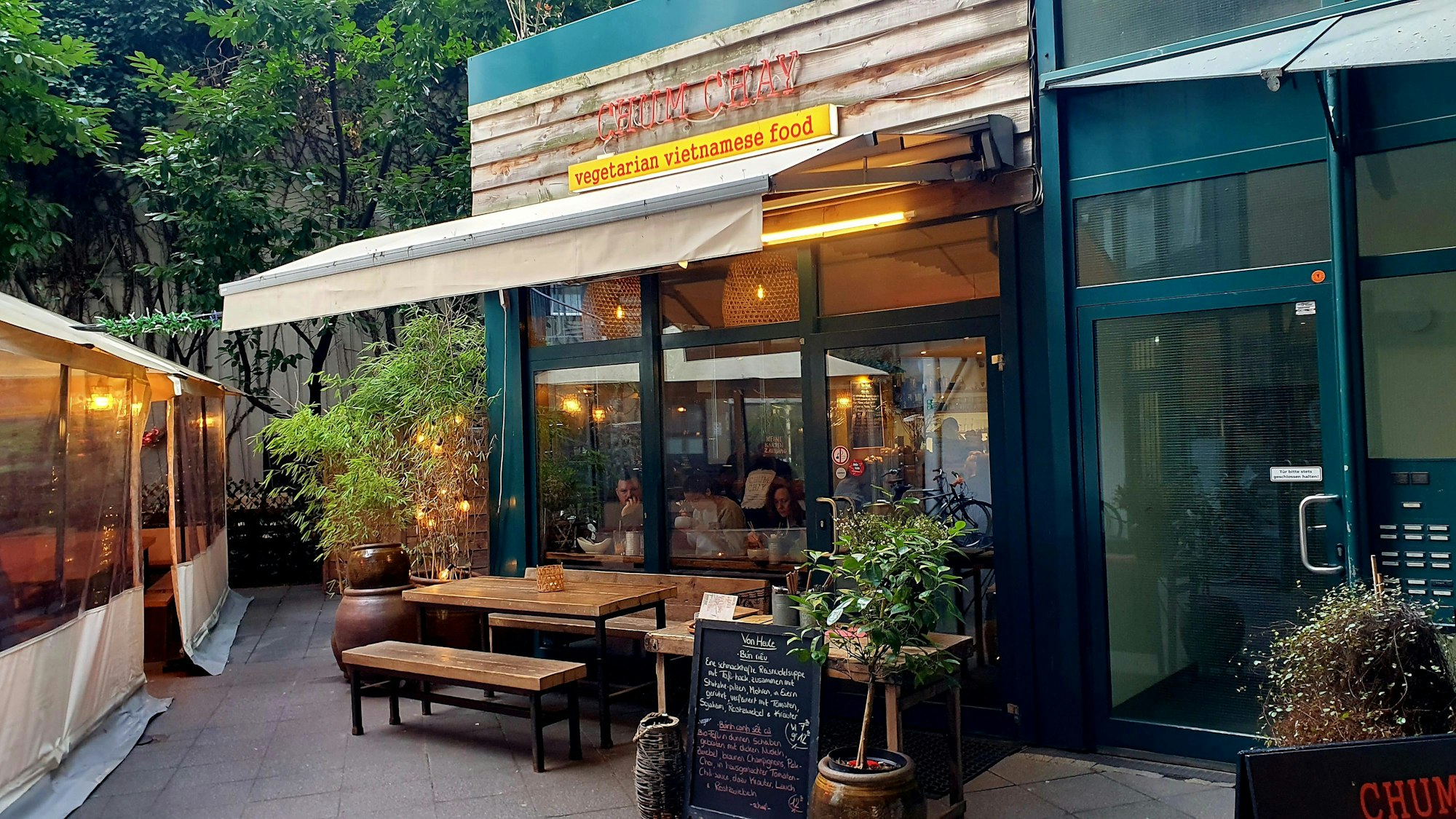 Die Fensterfront des Lokals ist von außen zu sehen, mit Markise und einigen Pflnzen davor.