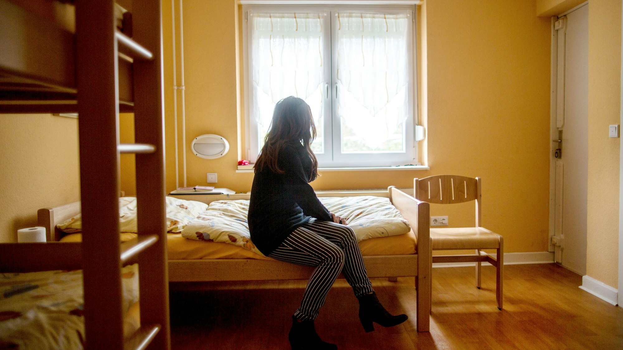 Eine Frau sitzt im Frauenhaus auf einem Bett. Das Gleichstellungsministerium in NRW will den Schutz von Frauen gegen Gewalt verbessern.