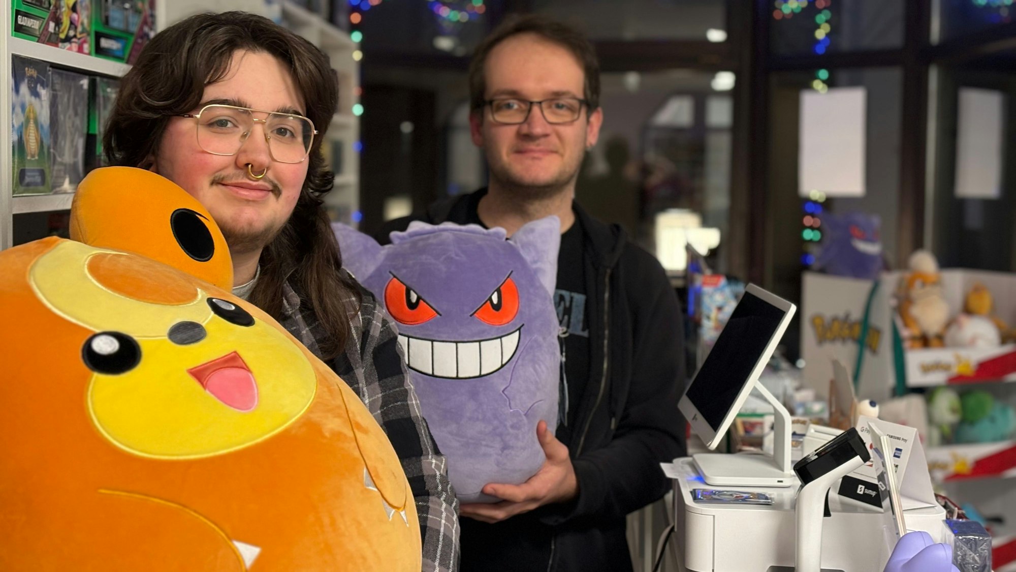 Zwei Männer mit Brillen halten Pokémon-Plüschfiguren in die Kamera.