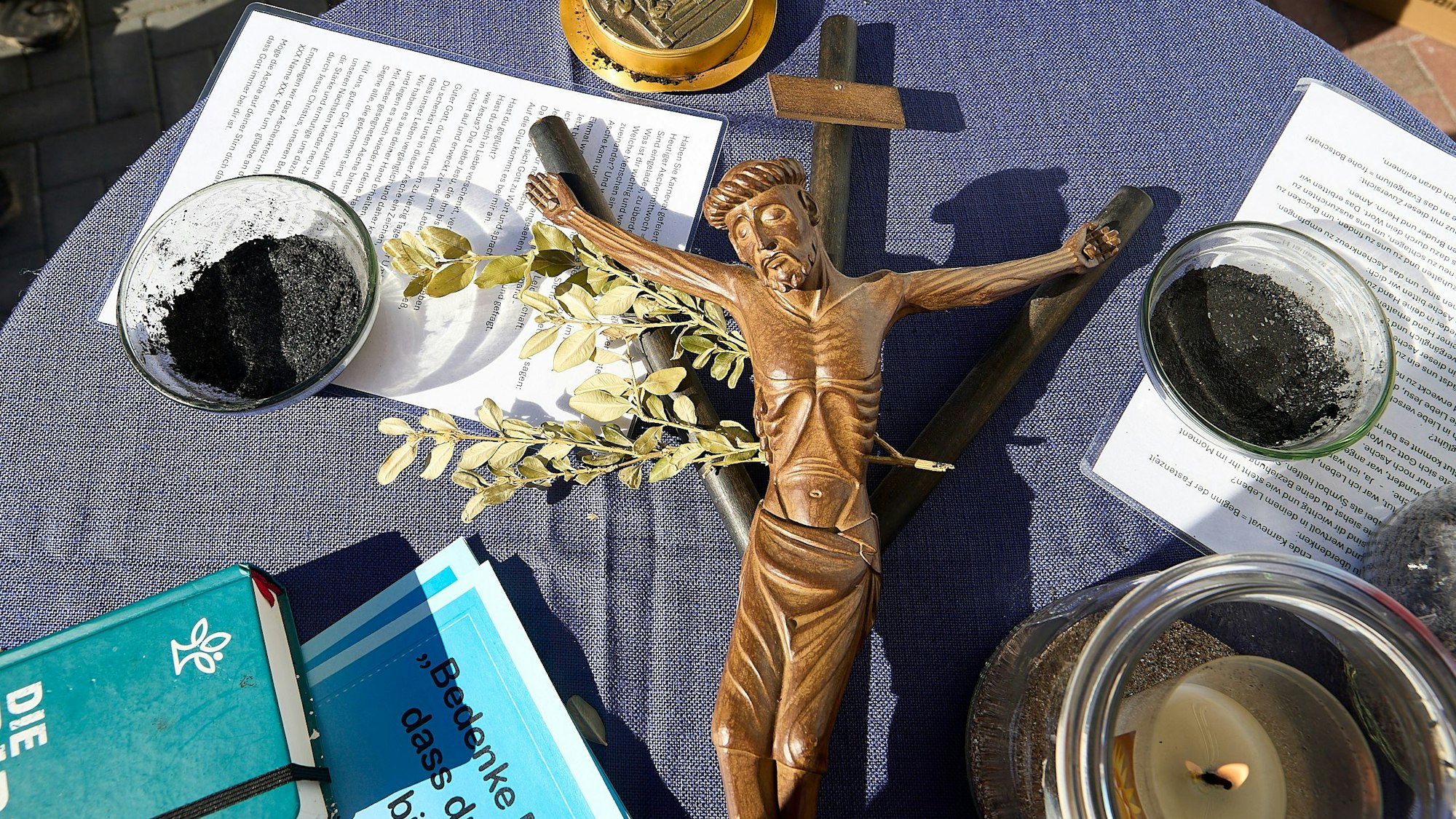 Auf einem Tisch sieht man ein Kruzifix, zwei Schälchen mit Asche, eine Kerze und Schriftstücke.