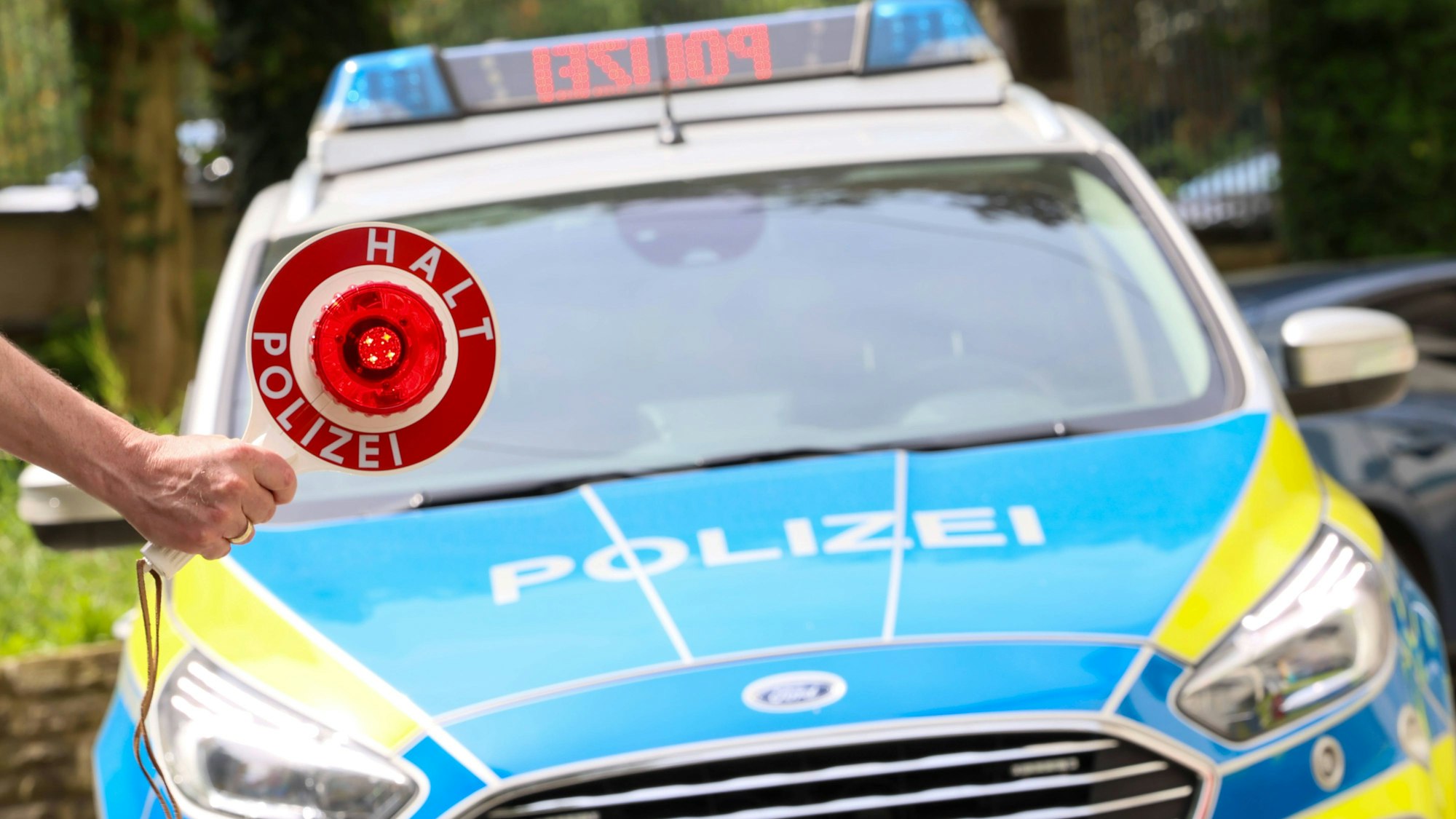 Ein Polizist hält eine Anhaltekelle vor einen Streifenwagen, auf dem Dachbalken steht Polizei.