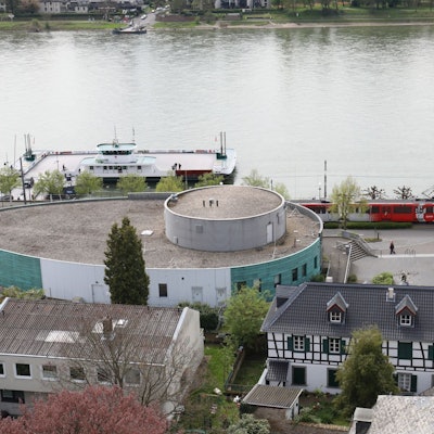 Ein ovales Gebäude mit grüner Fassade, dahinter der Rhein und die Fähre Königswinter.