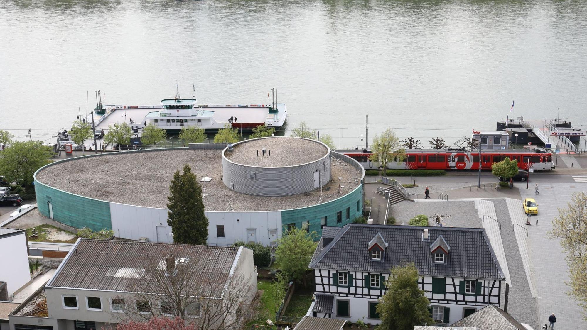 Ein ovales Gebäude mit grüner Fassade, dahinter der Rhein und die Fähre Königswinter.