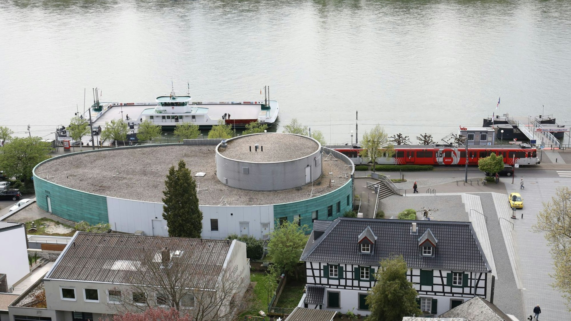 Ein ovales Gebäude mit grüner Fassade, dahinter der Rhein und die Fähre Königswinter.
