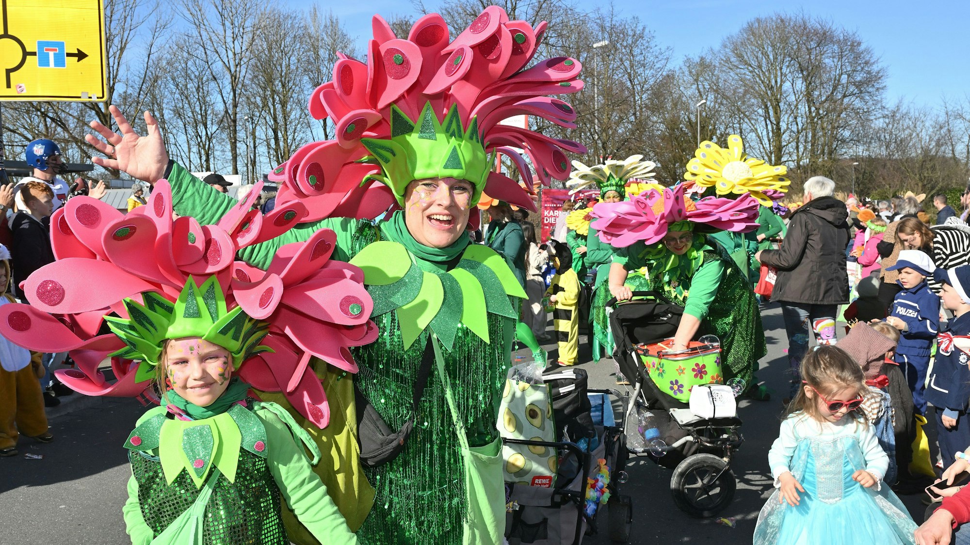 Ein Kind und sein Elternteil sind als glitzernde Blumen verkleidet und tragen grüne Kostüme und pinke Hüte. Im Hintergrund gucken viele Kostümierte den Kinderzug an.