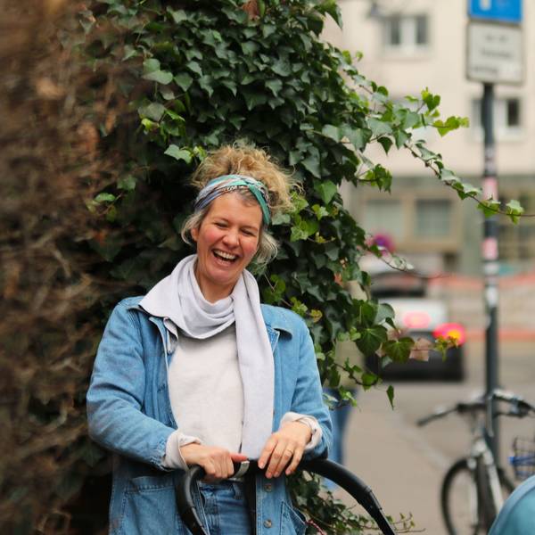 Einen Kinderwagen schiebend steht Susan Bollig vor einer Hecke auf einem Gehweg und lacht.