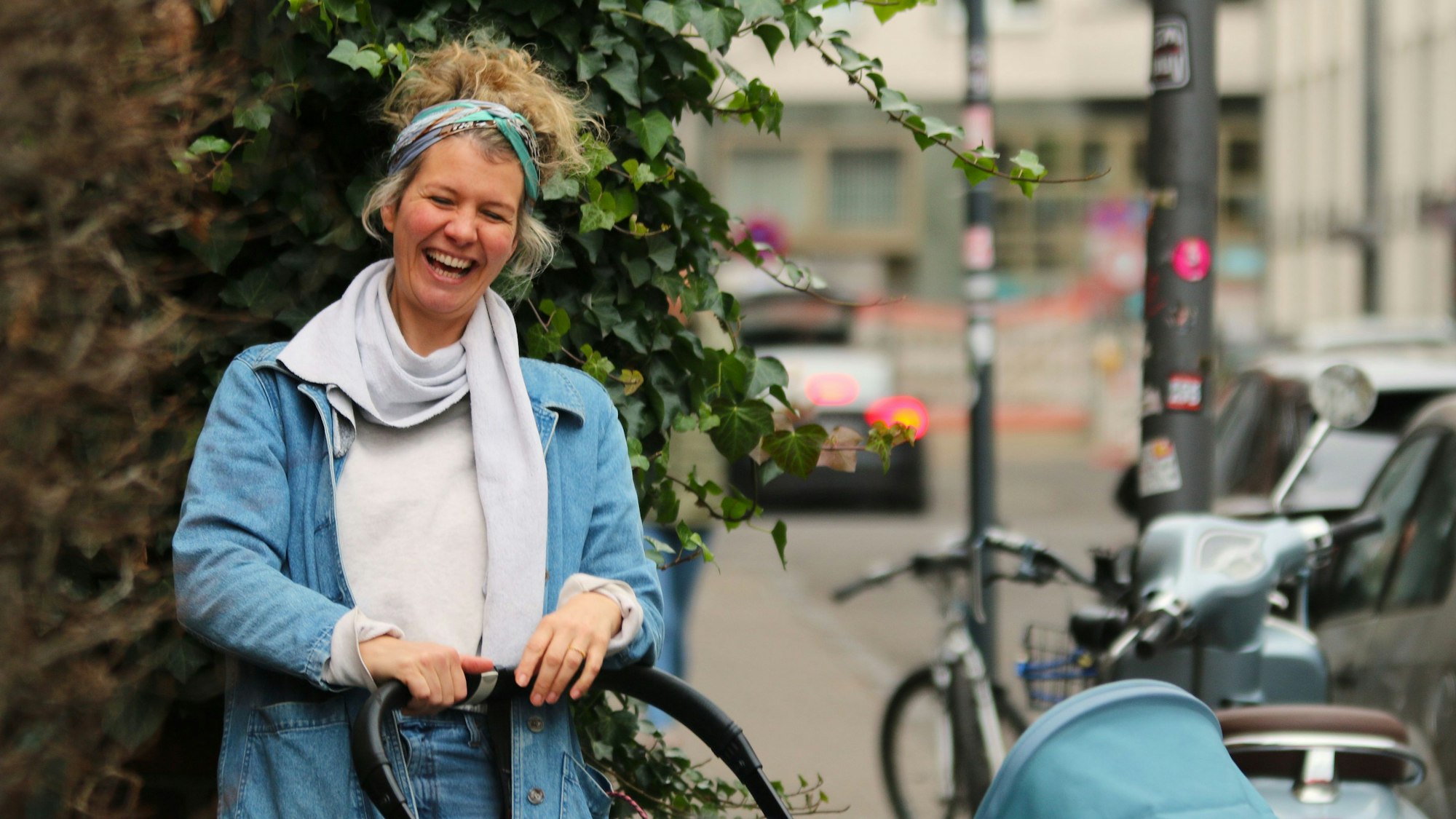 Einen Kinderwagen schiebend steht Susan Bollig vor einer Hecke auf einem Gehweg und lacht.