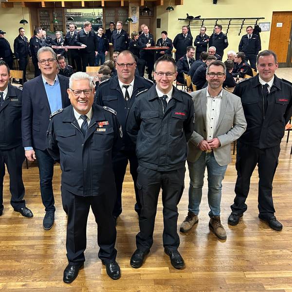 Fünf Männer in dunkelblauen Feuerwehruniformen und zwei in Zivilkleidung stehen im Vordergrund. Im Hintergrund sind weitere Feuerwehrleute zu sehen.