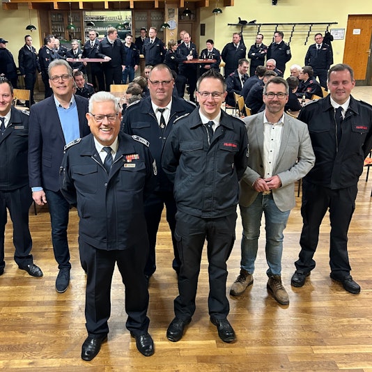 Fünf Männer in dunkelblauen Feuerwehruniformen und zwei in Zivilkleidung stehen im Vordergrund. Im Hintergrund sind weitere Feuerwehrleute zu sehen.