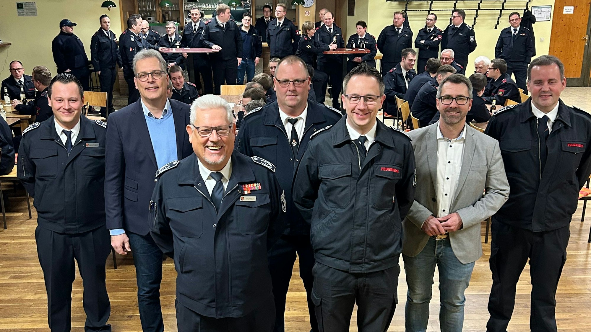 Fünf Männer in dunkelblauen Feuerwehruniformen und zwei in Zivilkleidung stehen im Vordergrund. Im Hintergrund sind weitere Feuerwehrleute zu sehen.