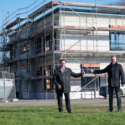 Das Foto zeigt zwei Männer vor dem eingerüsteten zweigeschossigen Rohbau.