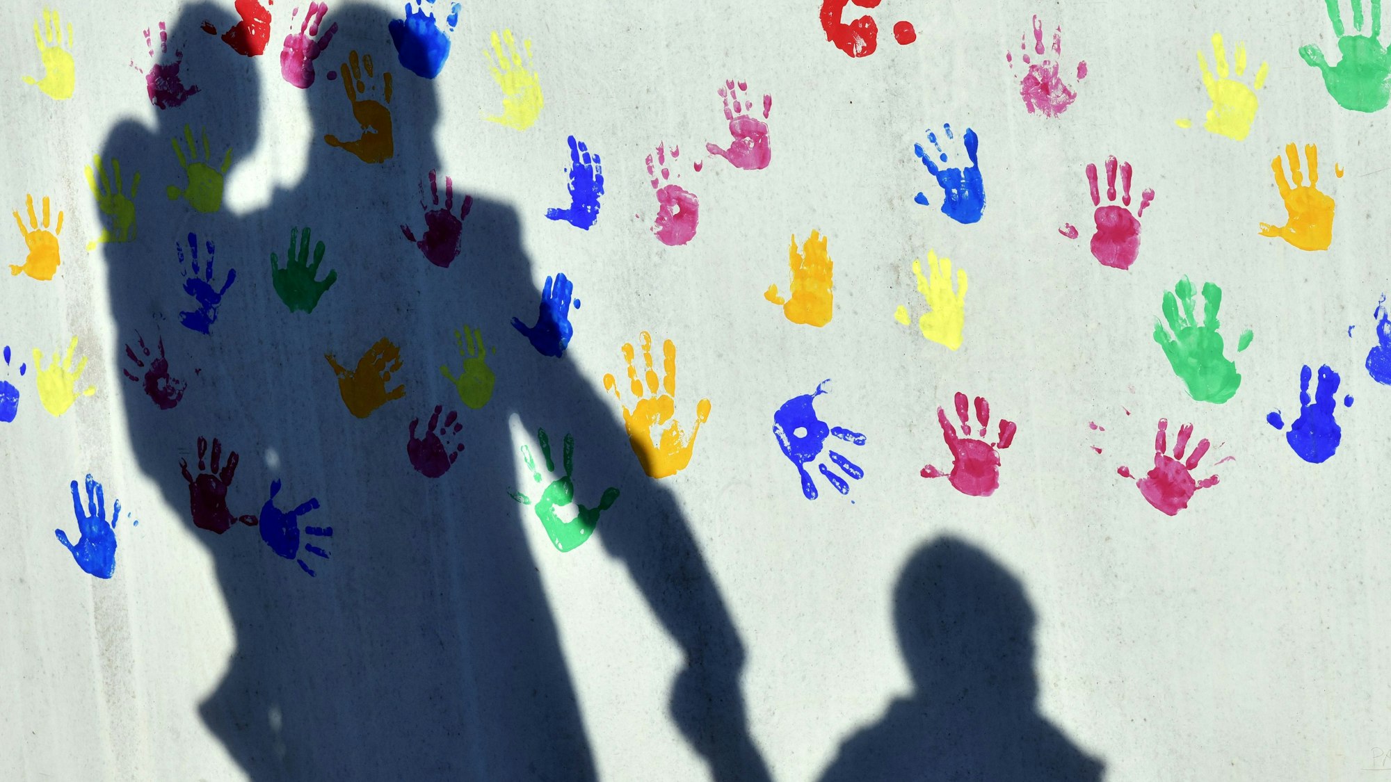 Ein Mann mit einem Kind auf dem Arm und einem an der Hand wirft einen Schatten auf eine mit bunten Handabdrücken bemalte Wand einer Kindertagesstätte.