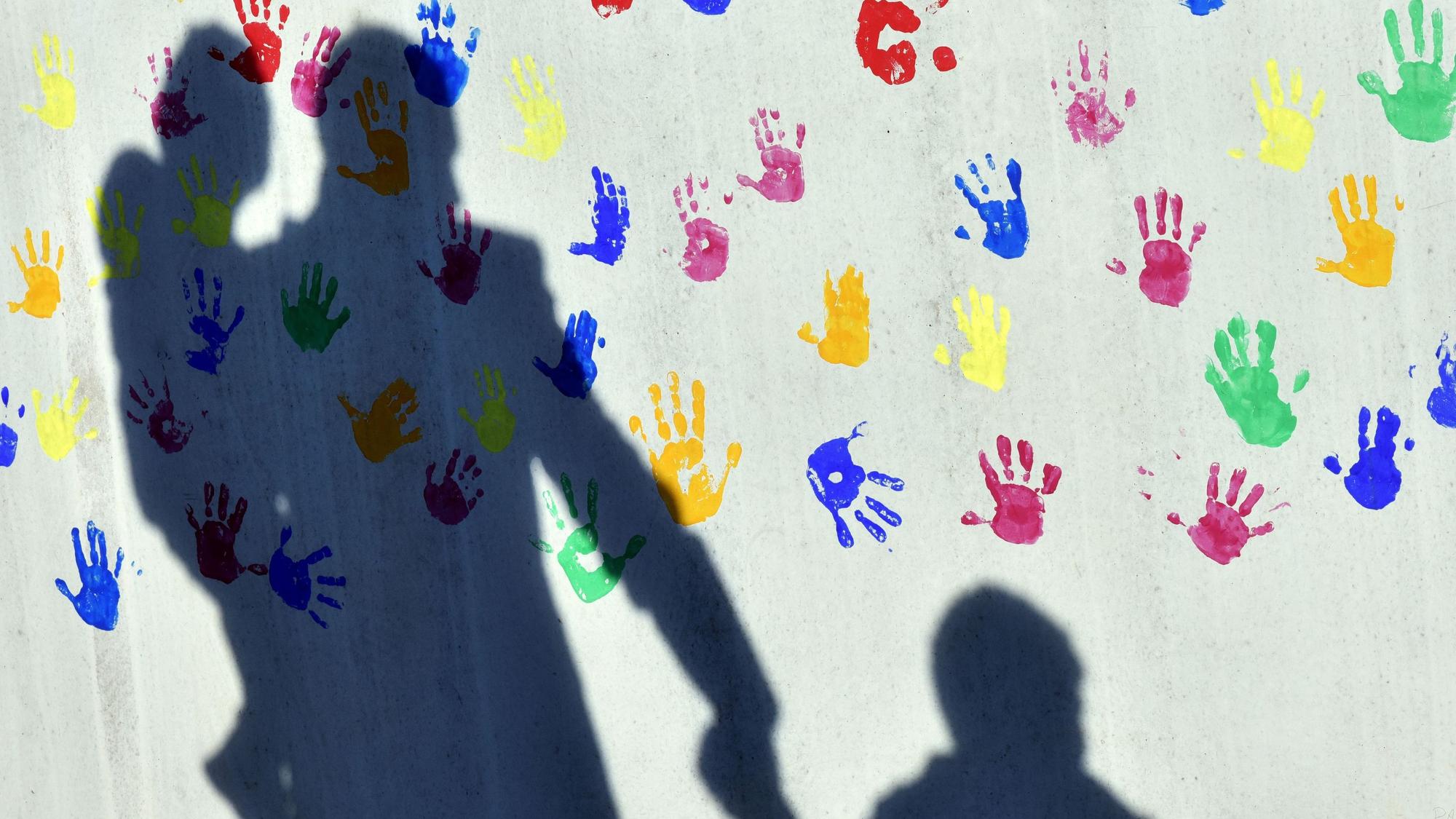 Ein Mann mit einem Kind auf dem Arm und einem an der Hand wirft einen Schatten auf eine mit bunten Handabdrücken bemalte Wand einer Kindertagesstätte.