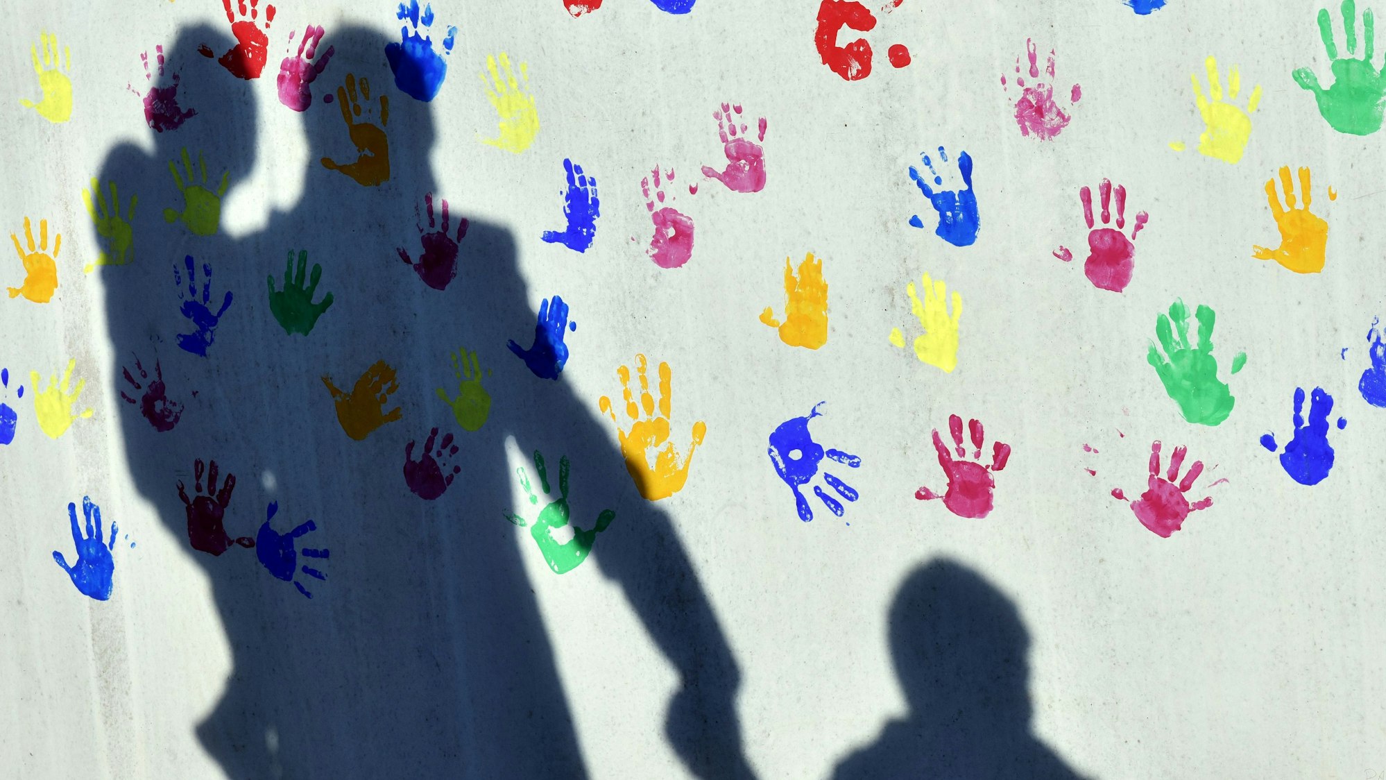 Ein Mann mit einem Kind auf dem Arm und einem an der Hand wirft einen Schatten auf eine mit bunten Handabdrücken bemalte Wand einer Kindertagesstätte.