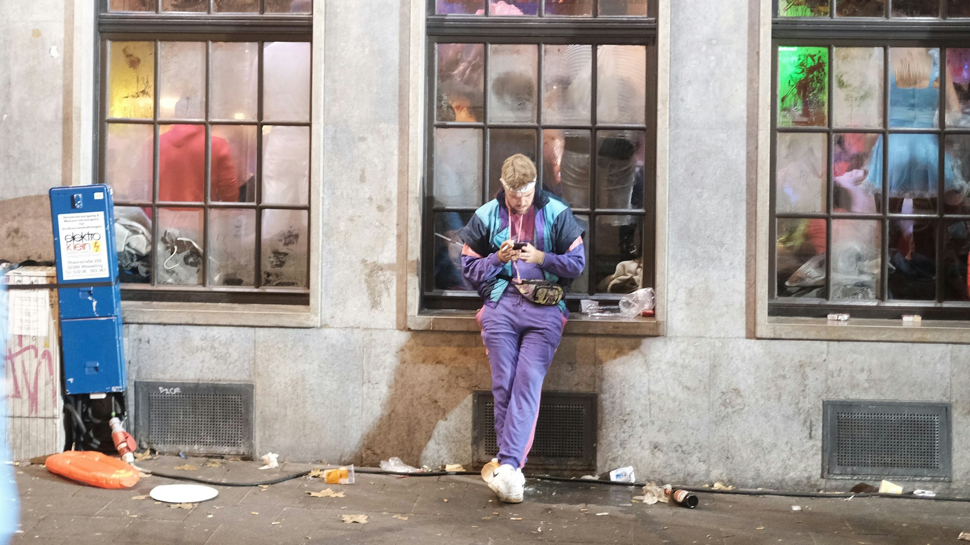 Ein junger Jeck beschäftigt sich mit seinem Handy, während hinter ihm in der Kneipe im Kwartier Latäng Karneval gefeiert wird.