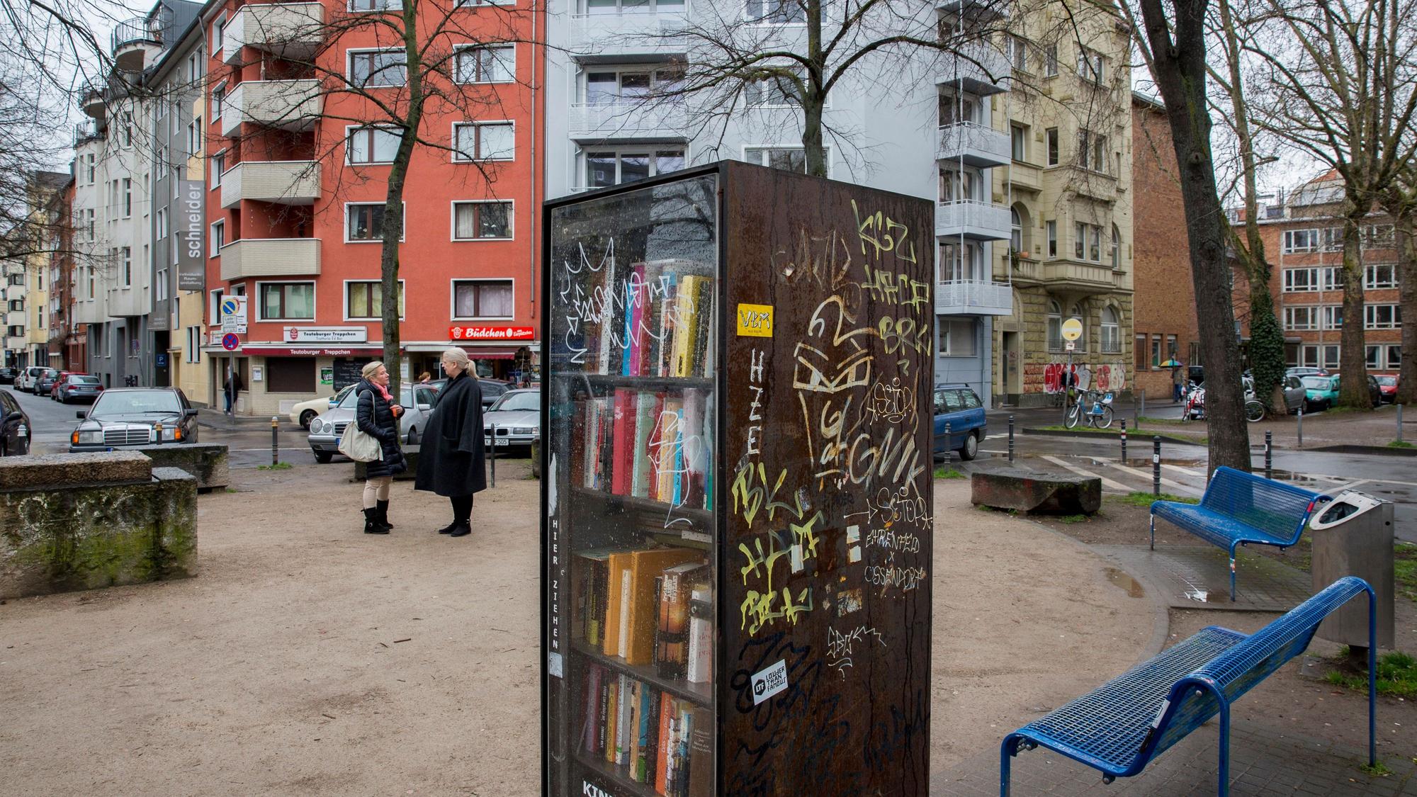 In der Mitte des von Wohnhäusern gesäumten, „Eierplätzchen“ genannten Platzes in der Kölner Südstadt steht ein Bücherschrank. An den Seiten des Platzes sind Sitzbänke aufgestellt. Zwei Frauen stehen auf dem Platz und unterhalten sich.