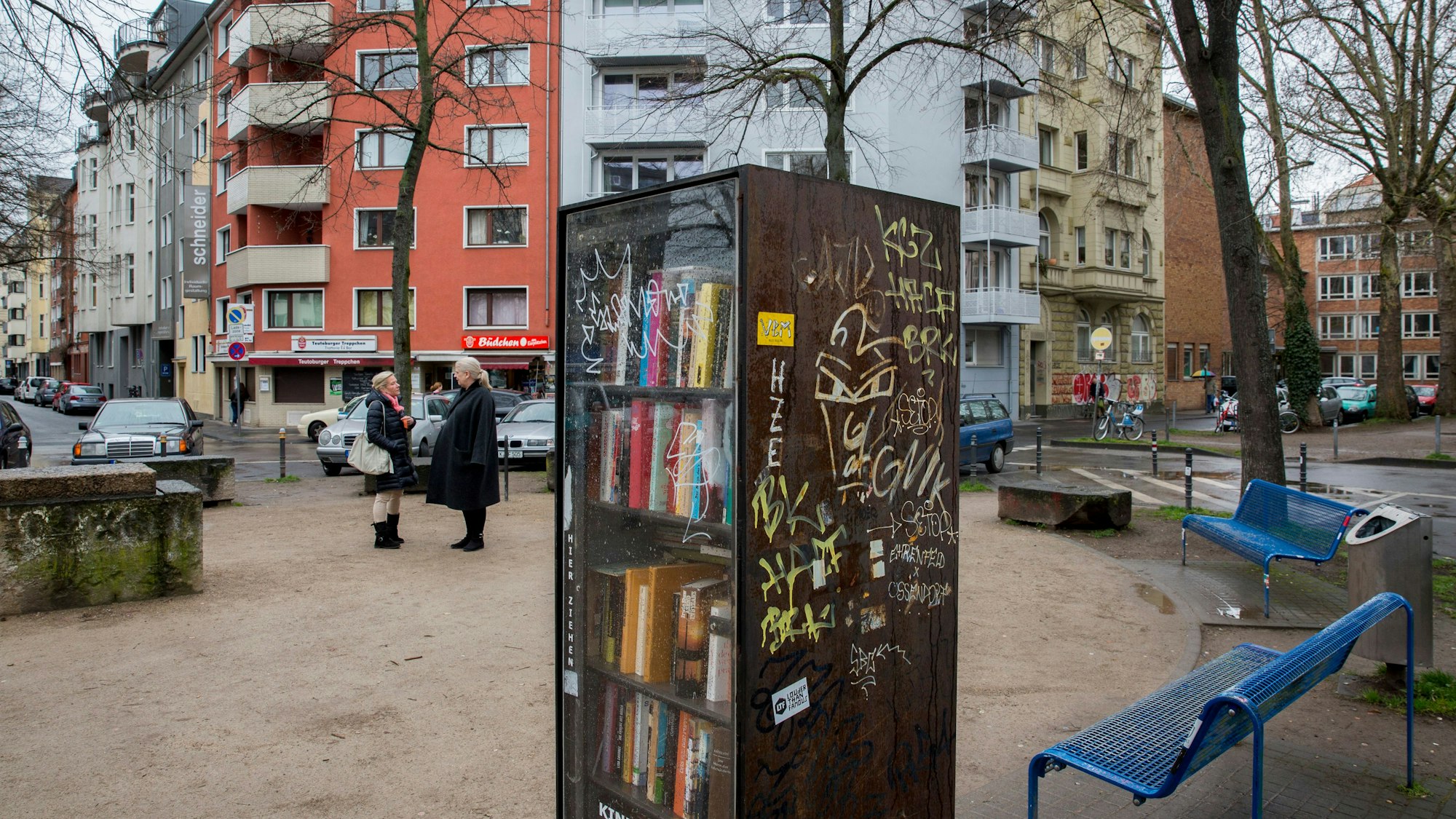 In der Mitte des von Wohnhäusern gesäumten, „Eierplätzchen“ genannten Platzes in der Kölner Südstadt steht ein Bücherschrank. An den Seiten des Platzes sind Sitzbänke aufgestellt. Zwei Frauen stehen auf dem Platz und unterhalten sich.