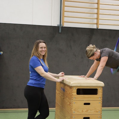 Eine Frau ist auf zwei Kasten-Turngeräte gestützt und lässt in Plank-Position einen Reifen Kreisen. Eine andere Frau steht lächelnd neben ihr.