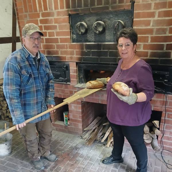 Ein Mann und eine Frau stehen an einem Steinbackofen. Er zieht auf einem Schieber ein Brot aus dem Ofen, sie hält eines in der Hand.