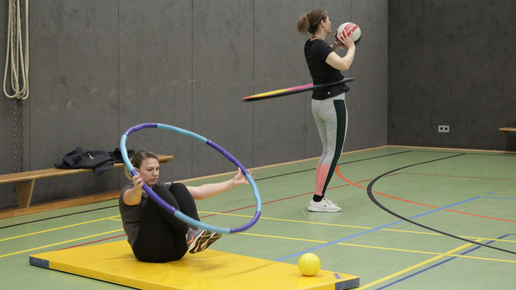 Eine Kursteilnehmerin macht Bauchmuskel-Übungen mit Reifen auf einer Matte, eine andere lässt den Hula Hoop Reifen kreisen und wirft einen Ball.