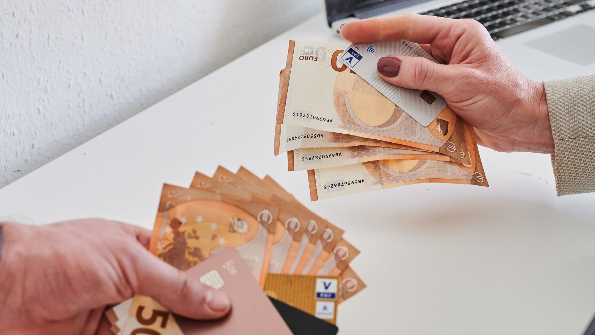 Das Bild zeigt eine männliche und eine weibliche Hand, die jeweils Geldscheine und Kreditkarten halten. Die männliche Hand hält mehr Geld und elektronische Zahlungsmittel in der Hand, die weibliche weniger.