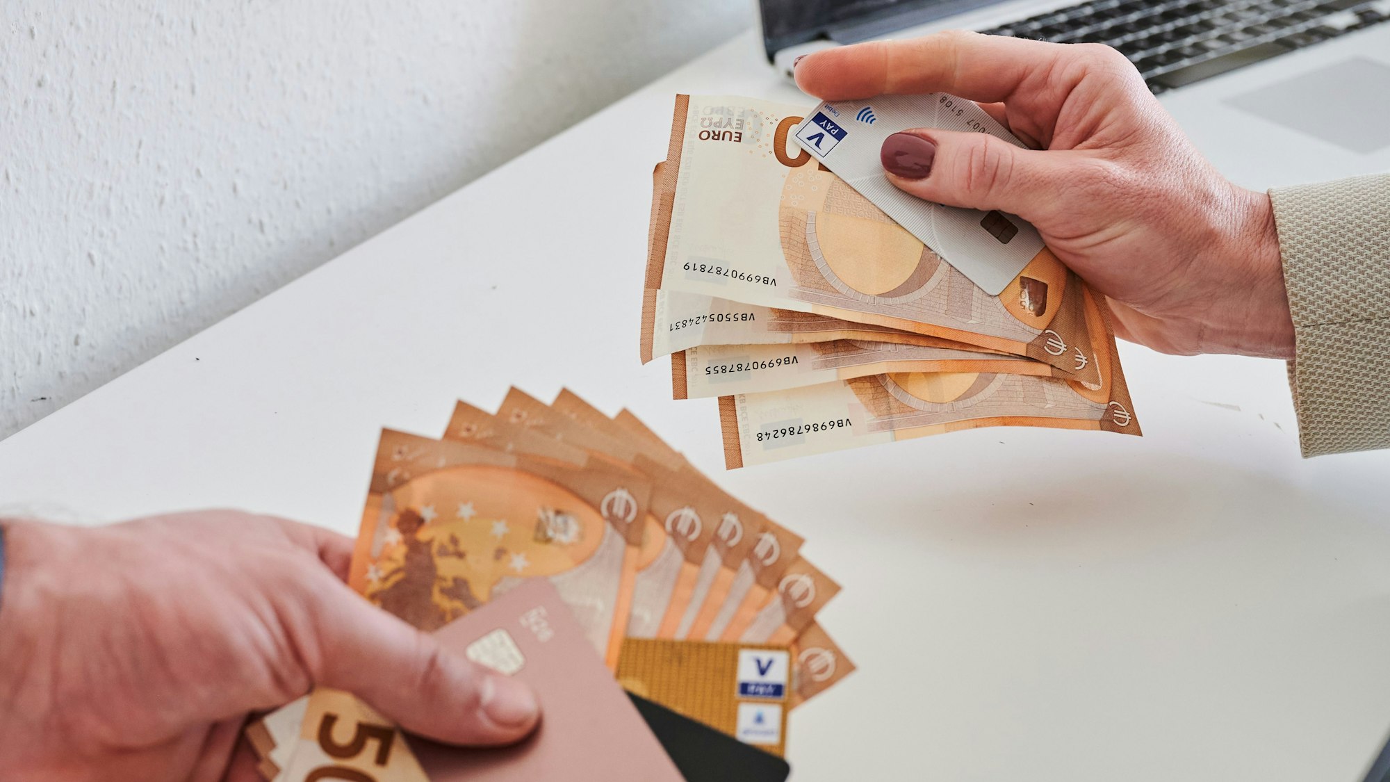 Das Bild zeigt eine männliche und eine weibliche Hand, die jeweils Geldscheine und Kreditkarten halten. Die männliche Hand hält mehr Geld und elektronische Zahlungsmittel in der Hand, die weibliche weniger.