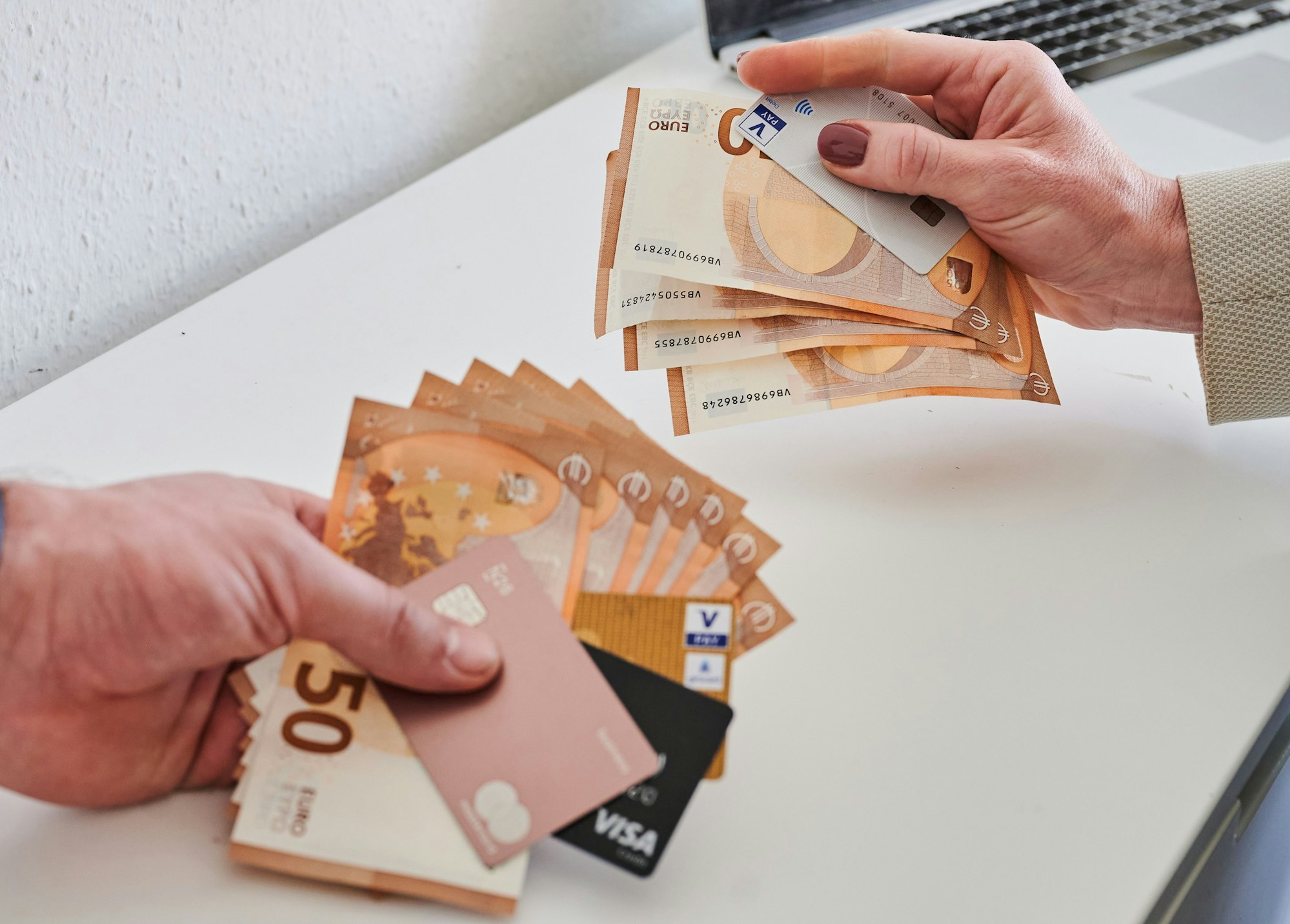 Das Bild zeigt eine männliche und eine weibliche Hand, die jeweils Geldscheine und Kreditkarten halten. Die männliche Hand hält mehr Geld und elektronische Zahlungsmittel in der Hand, die weibliche weniger.