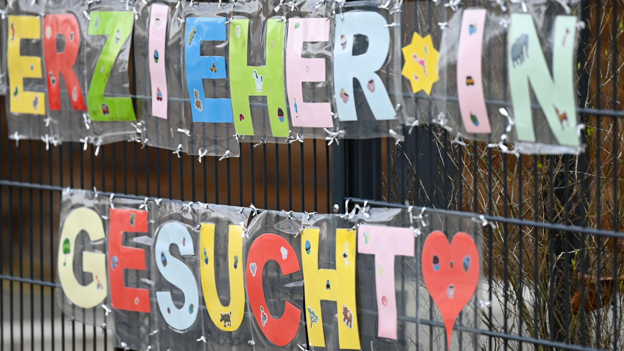 Mit bunten Buchstaben geformt sind die Worte „Erzieherin gesucht“ an einem Zaun einer Kindertagesstätte befestigt.