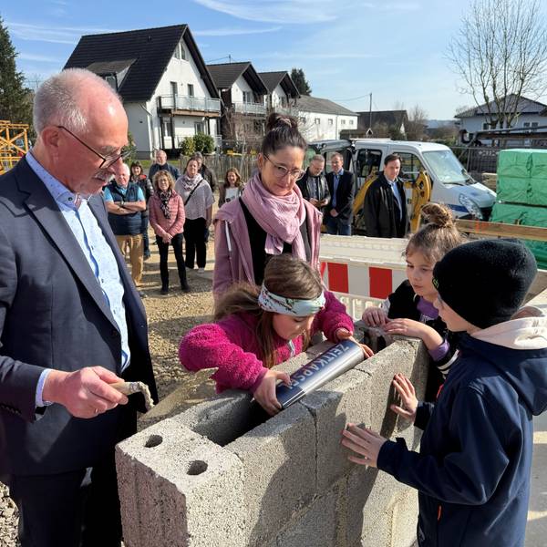 Ein Mann und eine Frau sehen zu, während Kinder eine Metallkapsel in einem Grundstein versenken.