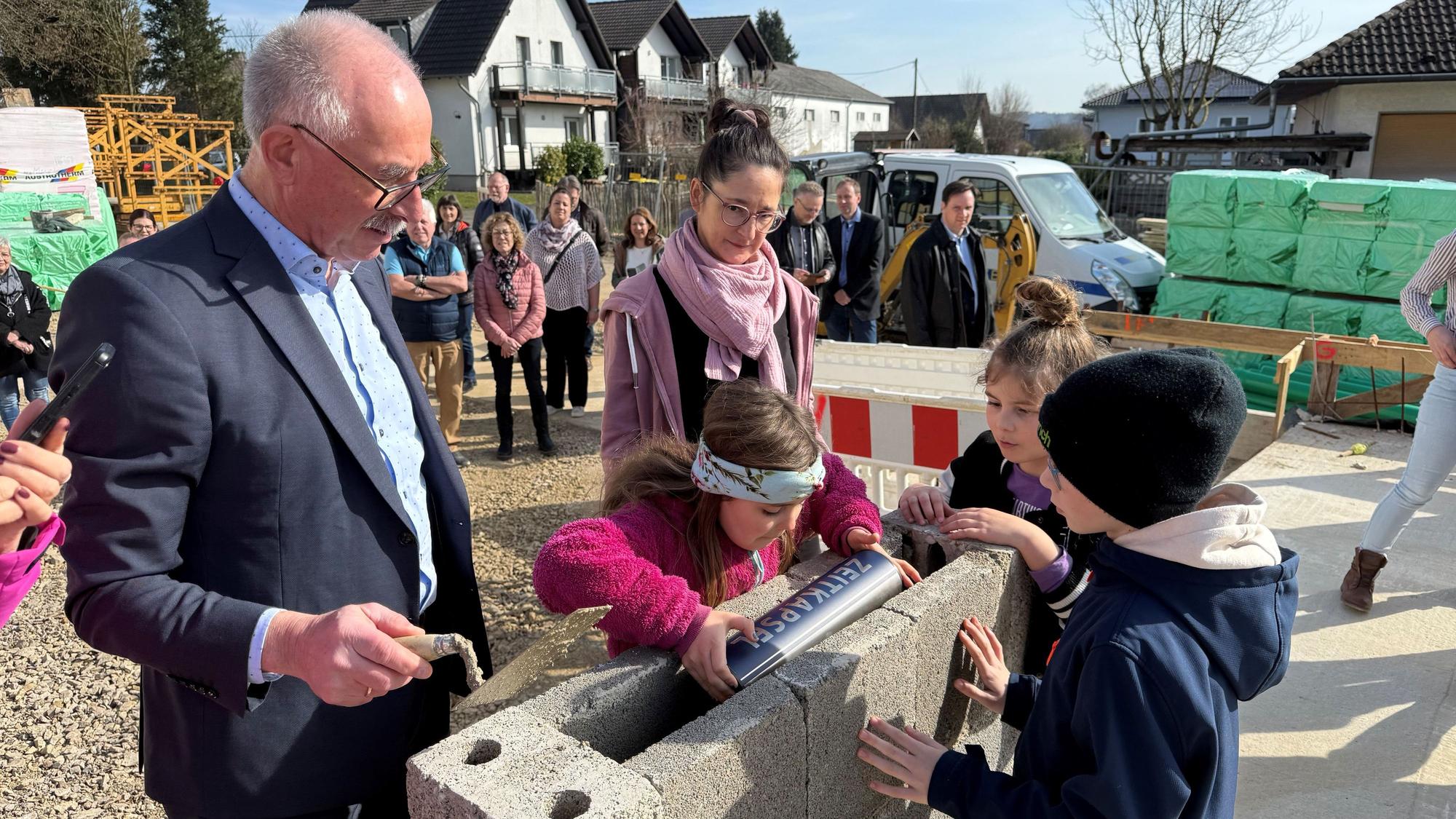 Ein Mann und eine Frau sehen zu, während Kinder eine Metallkapsel in einem Grundstein versenken.