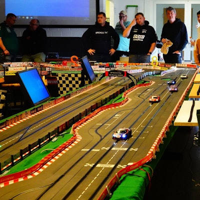 Auf dem Bild ist die Bahn zu sehen, die wie eine Rennstrecke auch zahlreiche Tafeln von mit Autos verbundenen Sponsoren zu sehen sind. Hinter der Bahn stehen die Teilnehmer des Rennens in einer Reihe.