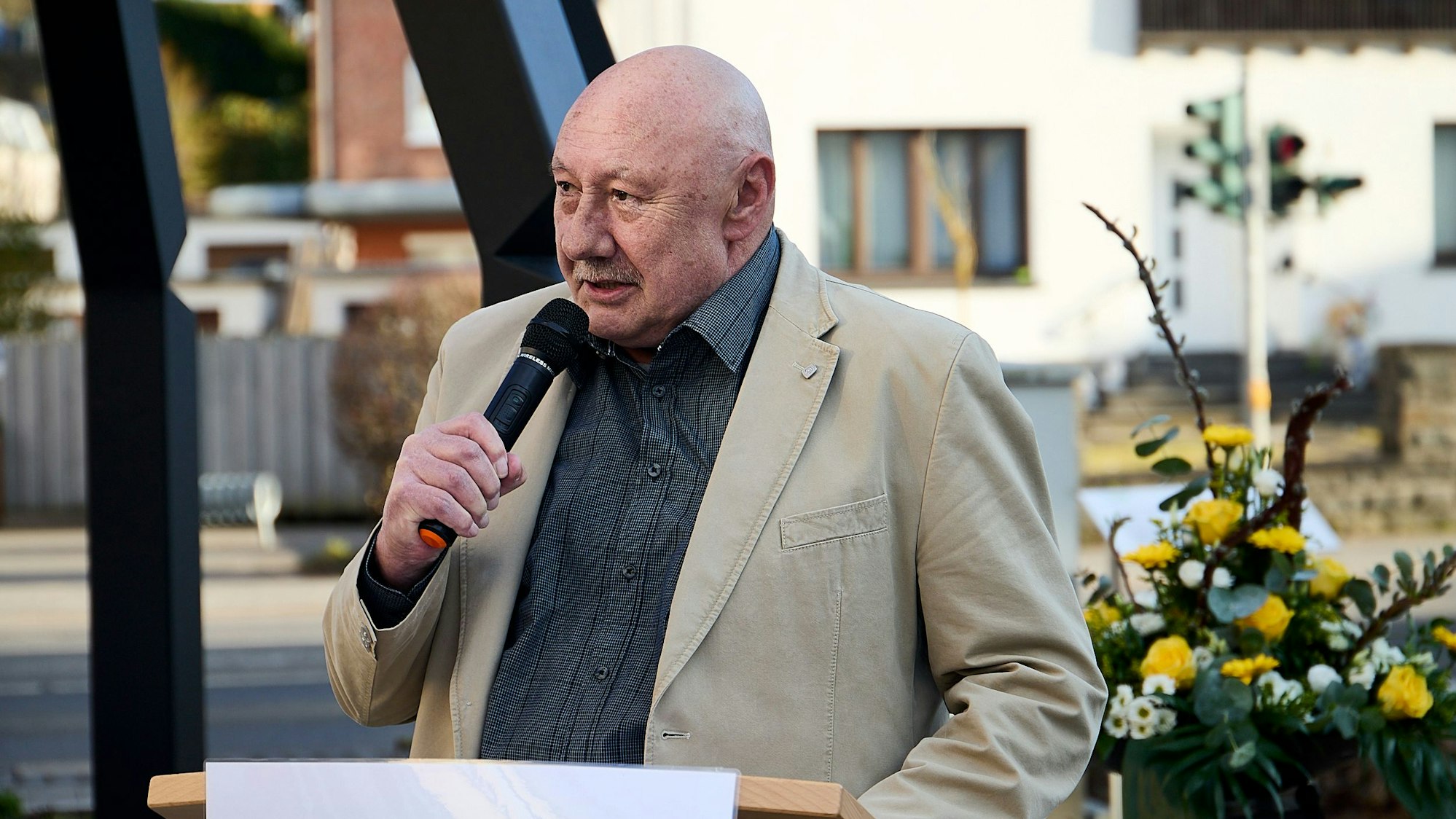 Der Journalist steht an einem Rednerpult. Er trägt ein dunkles Hemd und ein helles Sakko, im Hintergrund ist ein Blumengesteck zu sehen.