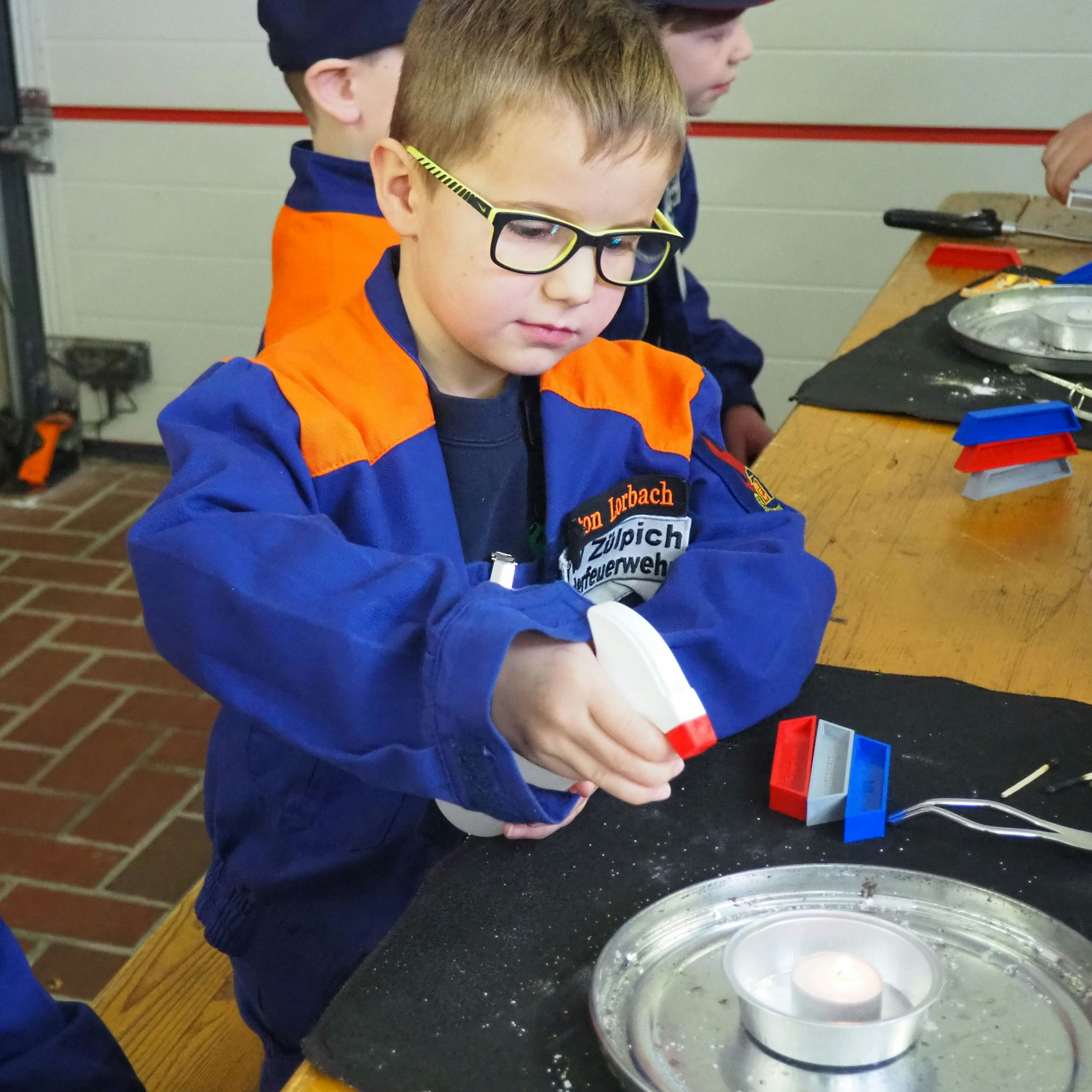 Ein Junge in orange-blauer Jacke sprüht mit einer Wassersprühflasche auf ein brennendes Teelicht.