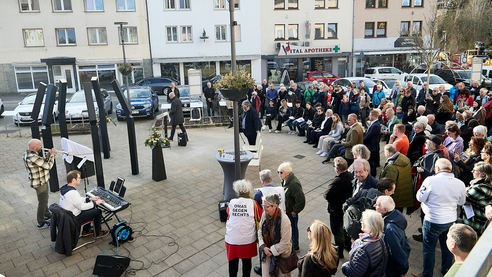 Eine Gruppe von Menschen steht auf einem Platz vor einer eingehüllten Tafel, die von sechs Metallstelen umgeben ist.