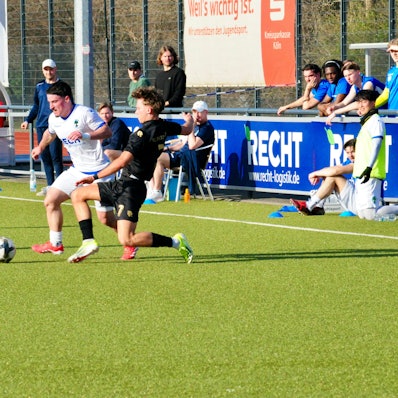 Das Foto zeigt einen Fußballer im weißen Trikot mit Ball am Fuß, der seinem Gegenspieler im schwarzen Dress bedrängt wird.