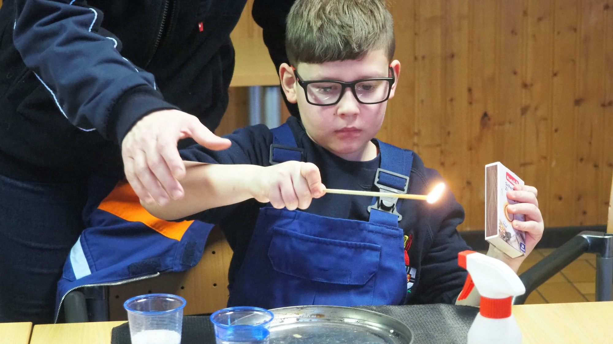 Ein Junge in blauer Latzhose und mit Brille hat ein langes brennendes Streichholz in der Hand.