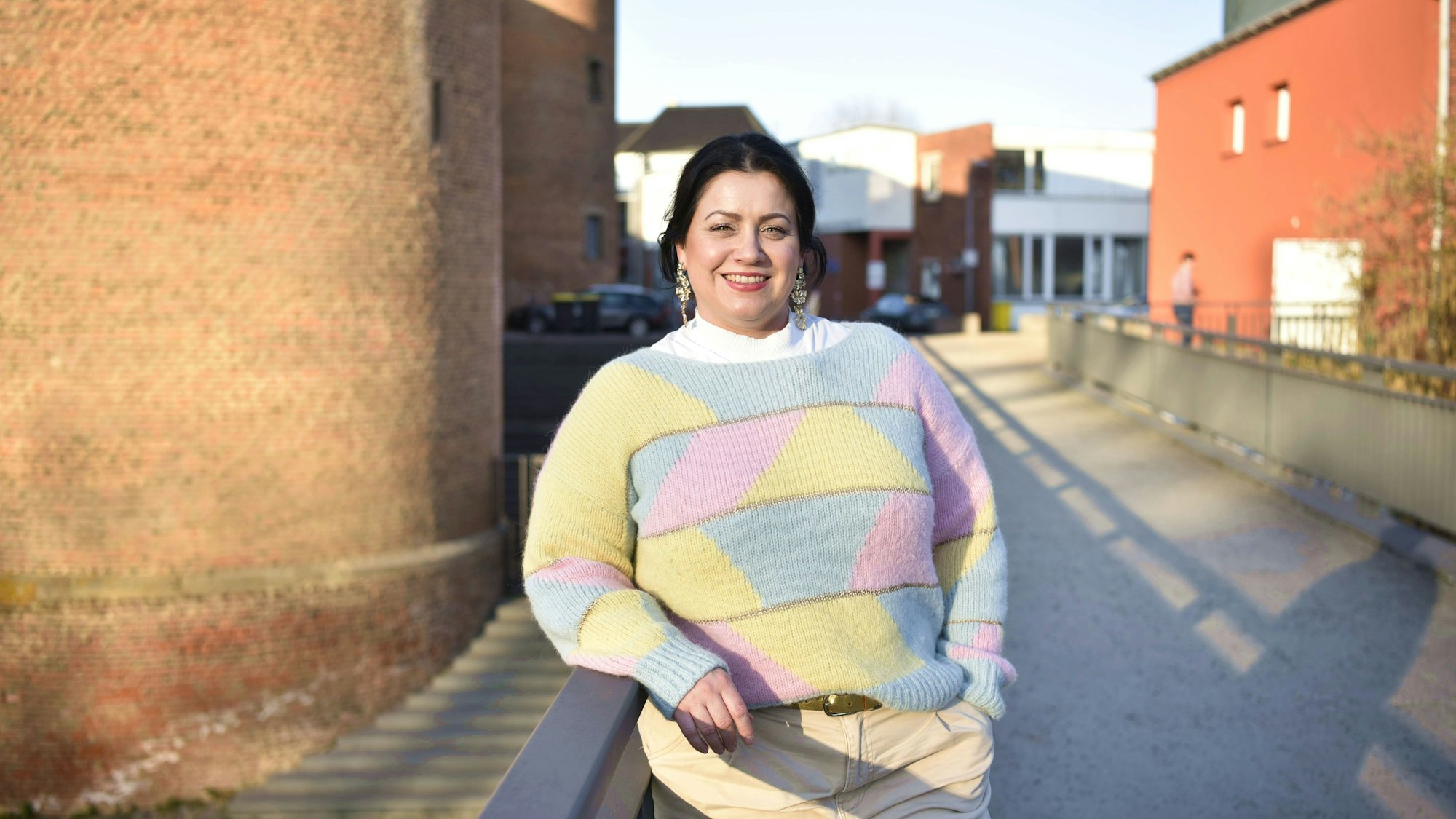 Die Kandidatin in pastellfarbenem Pullover steht an einem Geländer, im Hintergrund sind Mauern und Häuser zu erkennen.