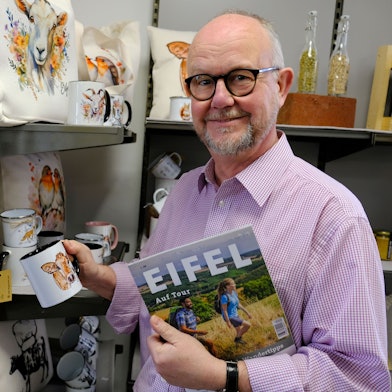 Der Schriftsteller Ralf Kramp steht vor einem Regal mit Eifel-Merch. Unter seinem linken Arm klemmt ein Tourismus-Magazin mit der Aufschrift „Eifel“.