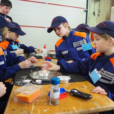 Kinder in orange-blauen Jacken sitzen an einem Tisch und experimentieren.