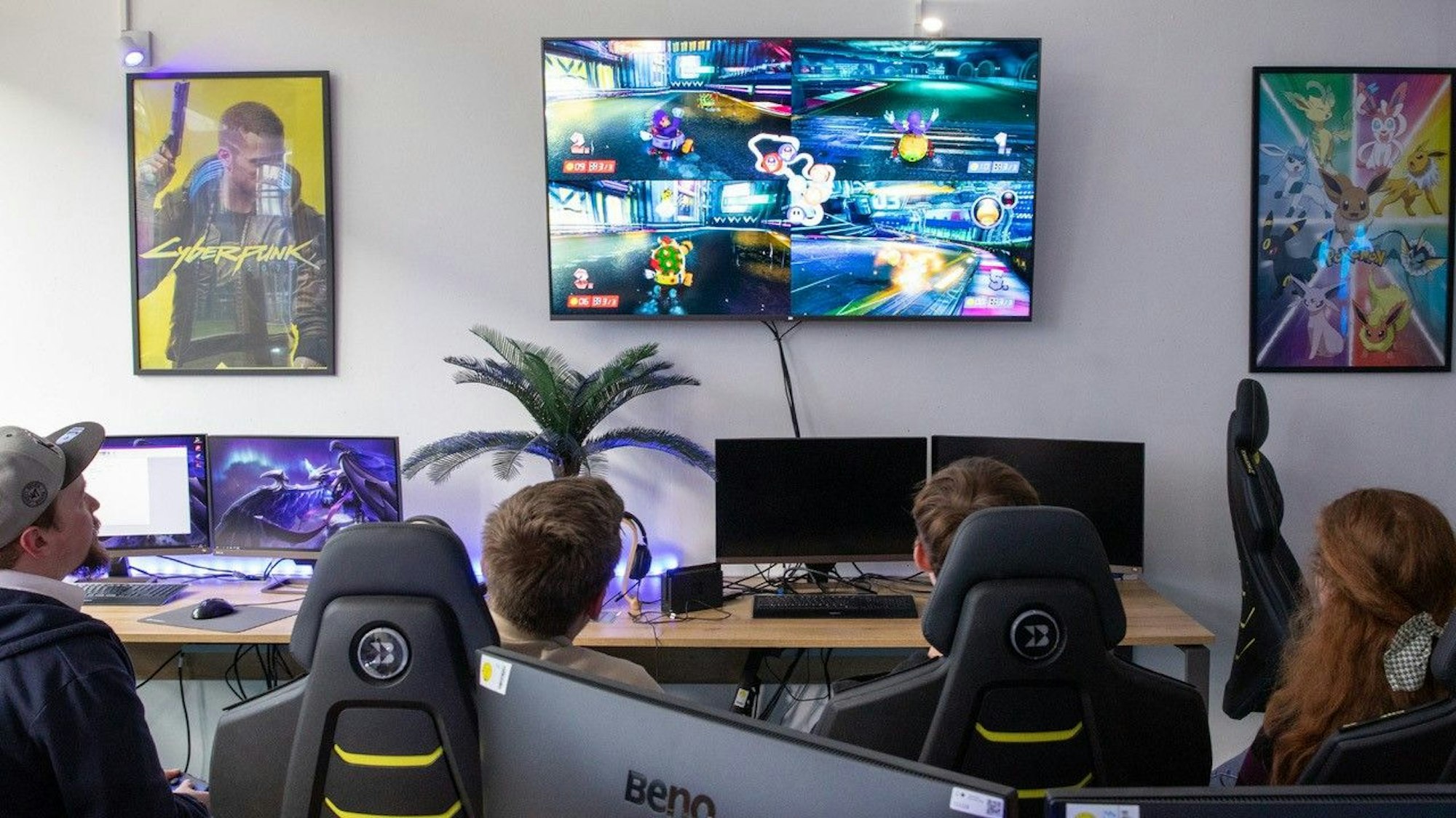 Junge Leute in speziellen Schreibtischstühlen sitzen vor einem großen Computerbildschirm, auf dem ein Spiel zu sehen ist.