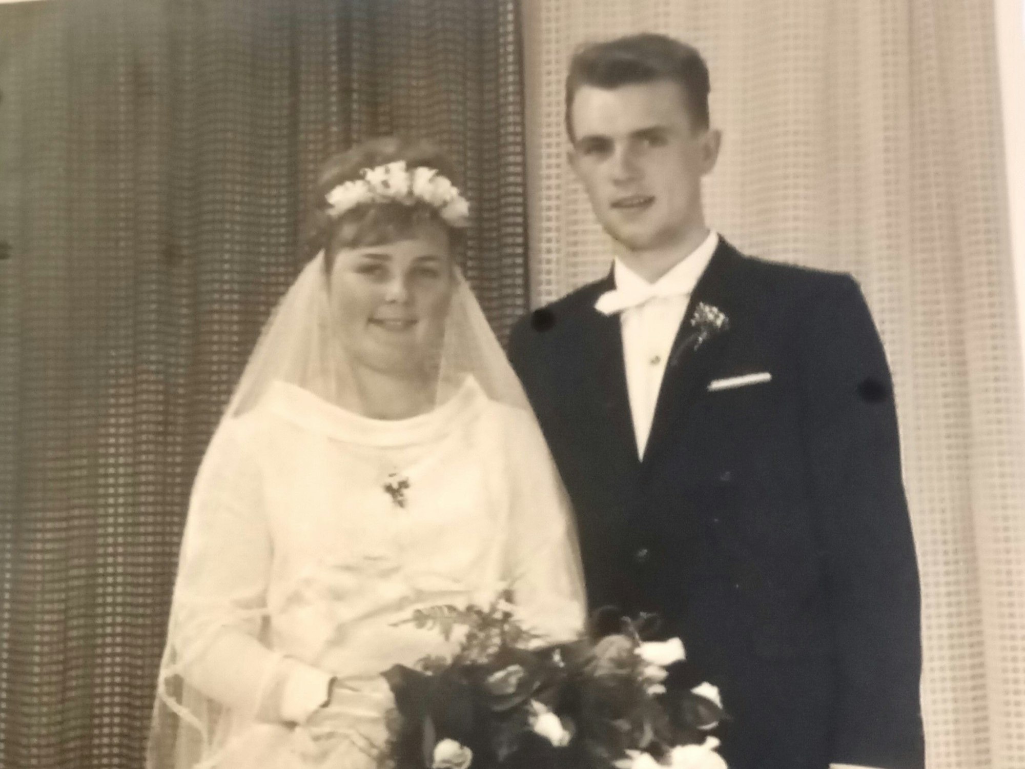 Die Braut trägt einen Schleier und hält Blumen in der Hand, der Bräutigam trägt einen Anzug. Das Foto ist schwarz-weiß.