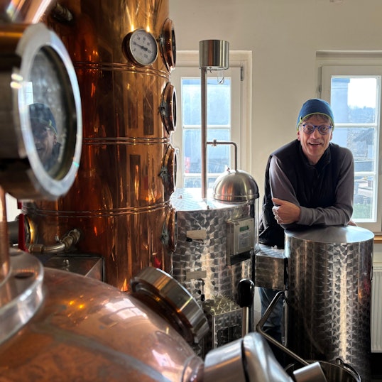 Ein Mann mit Kappe und Brille lehnt sich auf eine Destillationsanlage.