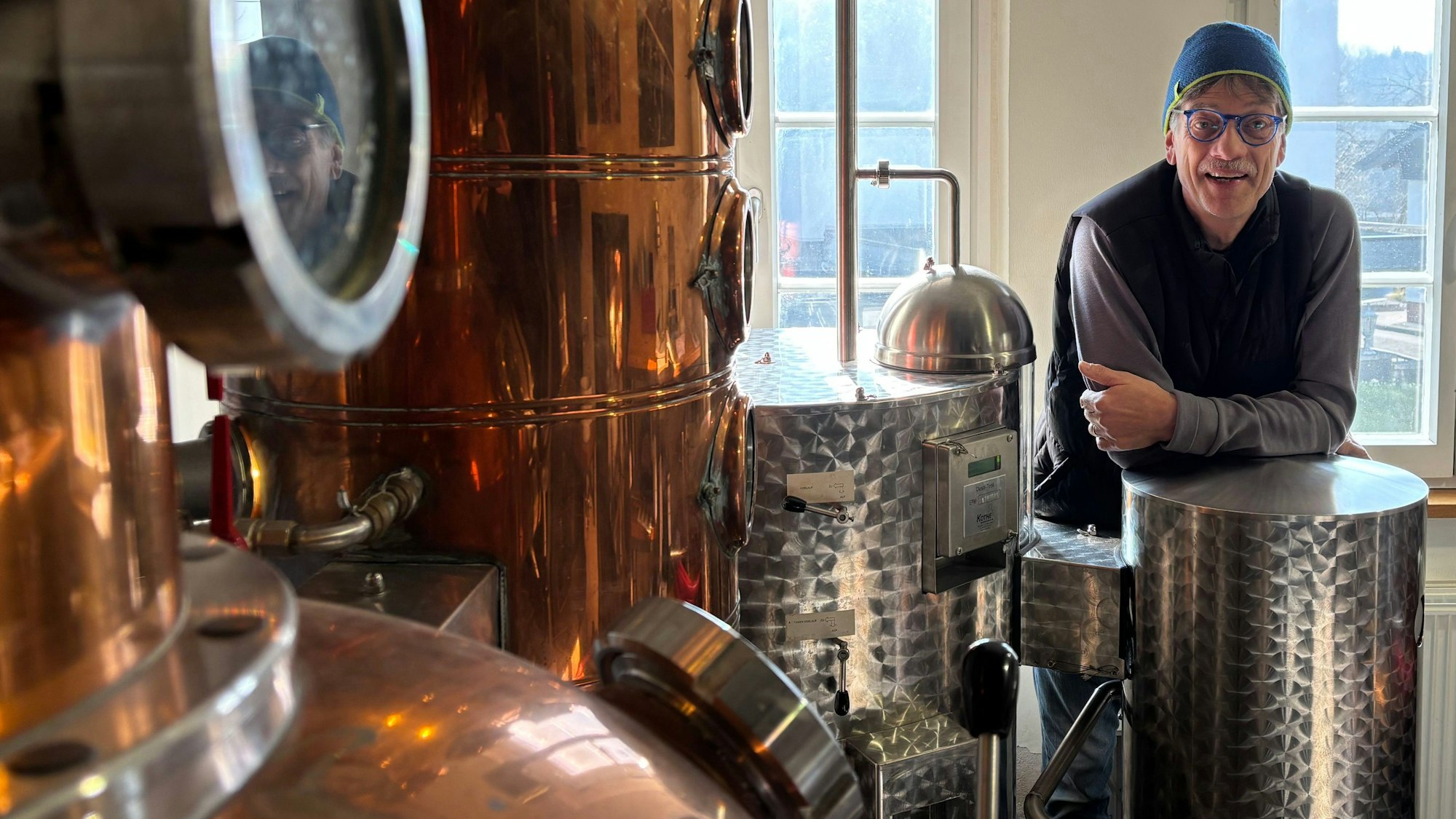 Ein Mann mit Kappe und Brille lehnt sich auf eine Destillationsanlage.