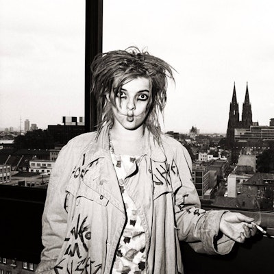 Nina Hagen raucht, den Kölner Dom im Hintergrund, eine Zigarette und zieht dabei die Wangen ein.
